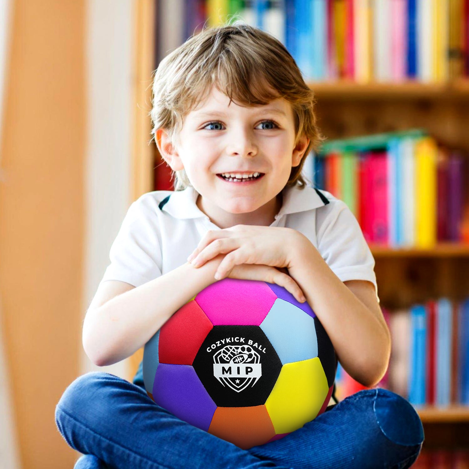 Colorful CozyKicks soccer ball held by smiling boy in a white polo shirt, sitting in front of a bookshelf.