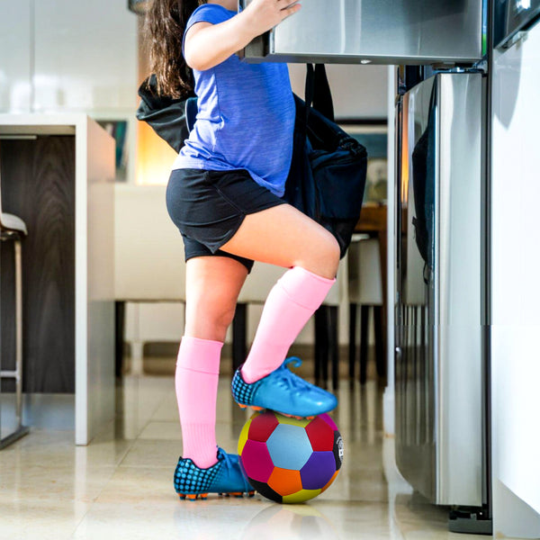 Child in blue athletic gear and pink socks stepping on a colorful soccer ball in a modern kitchen.