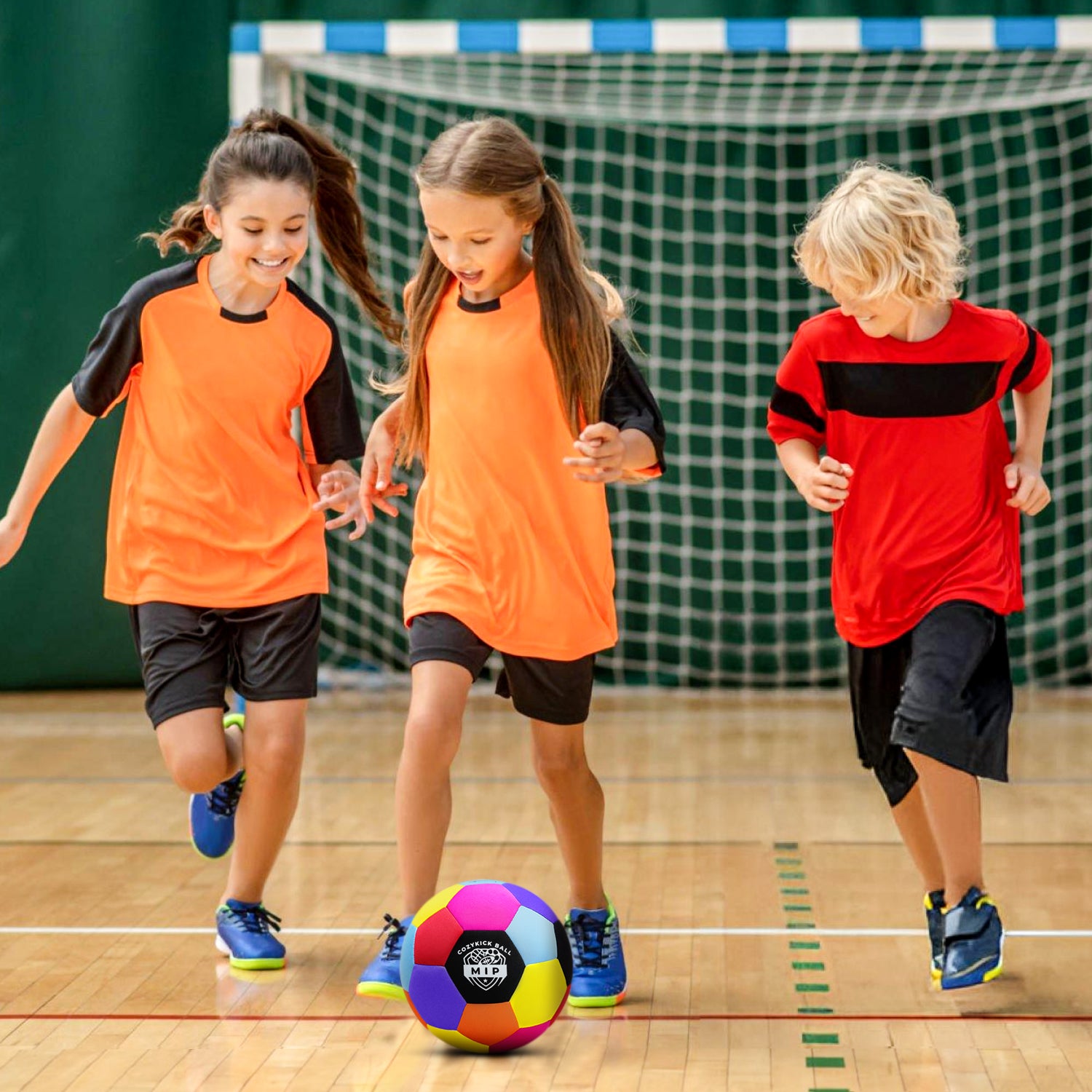Three children in orange and red athletic jerseys running towards a colorful soccer ball on a gym floor